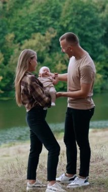 Beautiful Caucasian family of three standing in the nature at the river bank. Parents of newborn look at their child and then kiss. Vertical video
