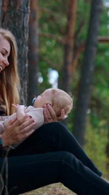 Happy first time mom speaking with her infant. Woman sits in the forest leaning on the tree and looking at her baby son. Vertical video