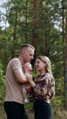Caucasian young family showing love to each other. Mother kisses her baby and man kisses his wife on the forehead. Nature at backdrop. Vertical video