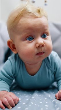 Adorable blue-eyed blond baby lies in the crib. Cute calm infant boy looking surprised around. Close up. Vertical video