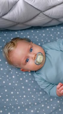 Lovely plump baby boy with pacifier lies in the crib holding legs up. Kid looks on the cot mobile hanging above. Top view. Vertical video