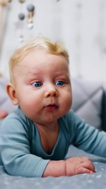 Blond Caucasian infant boy in blue bodysuit lies on the belly in the crib. Adorable blue-eyed boy looking up with surprise. Vertical video