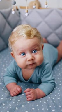 Caucasian infant with adorable plump cheeks lies peacefully in the crib. Lovely cute blue-eyed baby boy chewing something. Vertical video