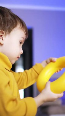 Dark-haired Caucasian toddler in yellow sweater turns a ring from pyramid in hands. Side view. Baby boy having fun at home. Low angle view. Vertical video