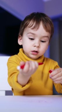 Beautiful Caucasian child sitting at desk. Baby boy dealing with a felt pen and pen cap. Low angle view. Vertical video