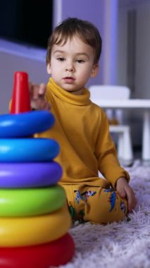 Cute Caucasian toddler in yellow pajama sitting on the fluffy plaid. Little boy plays with toy pyramid. Vertical video