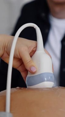 Female hand of a doctor holds the ultrasonic device on round belly of pregnant. Cropped image of man sitting at backdrop. Vertical video