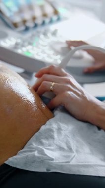 Pregnant woman is examined on ultrasonic equipment. Female doctor's hand moves the device by the big gelled belly of a future mom. Baby is kicking. Close up. Vertical video