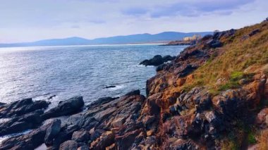 Rocks and boulders at the shore of the sea. Drone footage above the rocky coastline. A building at backdrop. Mountain range silhouette at backdrop.