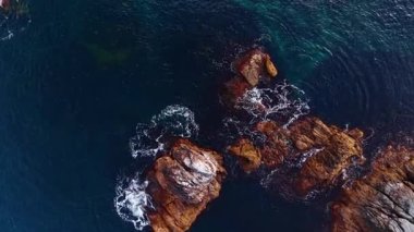 Spectacular deep blue waterscape splashes by the boulders sticking out of water. Top view on the rocky coast of the sea.