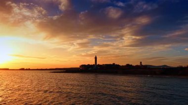 Dark silhouette of the skyline with a light house. Footage from above the seascape. Spectacular setting sun lights the sky, clouds and waterscape.
