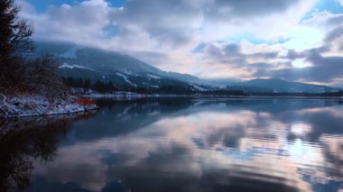 Dramatik kümülüs bulutları gölün huzurlu sularını yansıtır. Rıhtımdaki kayalıklarda kar var. Kış mevsimi doğası.