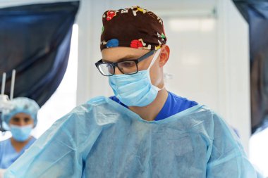 Ameliyat sırasında cerrah odaklandı. Koruyucu giysili bir cerrah iyi aydınlatılmış bir ameliyathanede dikkatle çalışır..