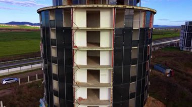 Silindir şeklinde yüksek katlı bir bina inşa ediliyor. Arkaplanda otoyolda nakliye araçları var. Hava perspektifi.