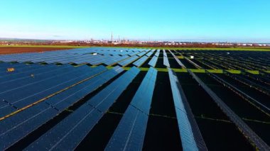 Modern güneş panellerinin uzun sıralarında hızlı bir uçuş. Kırsal kesimde fotovoltaik hücreleri olan yeşil alanlar. Rafineri fabrikası arkaplanda.