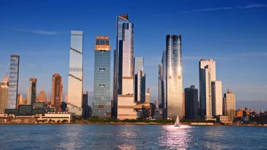 New York, ABD, 1 Ağustos 2025: Güneş ışığını yansıtan cam gökdelenler. Rıhtımdaki gemi yapısı. Bir yelkenli kıyıya yakın hareket ediyor. Nehir manzarasından görüntüle.