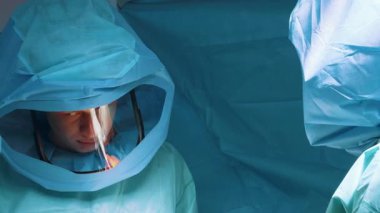 Mavi başlıklı iki cerrah yüksek konsantrasyonla ameliyat alanına yaslanıyor. Mavi örtülerle çevrili yoğun cerrahi prosedür sırasında tıbbi uzmanların yakın çekimi..