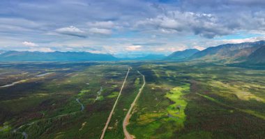 Tundra 'nın karşısındaki otoban ve demiryolu panoramik manzarası. Geniş, yüksek bir manzarada ıssız bir otoyol ve paralel tren yolu görünüyor. Uçsuz bucaksız, koyu yeşil Alaska tundrasını geçiyor.
