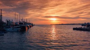 Bulutlu gökyüzünde batan güneş, Karadeniz 'in engebeli sularına yansıyor. Rıhtımda çok çeşitli tekneler var..
