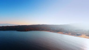 Haze, kıyıda bazı evler bulunan ormanlık kıyı şeridini kaplıyor. Sahil şehrinin sakin ve sakin bir manzarası. Hava görünümü.