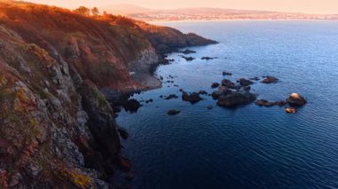 Rocky Coastal Cliff, Sunset Over Blue Ocean 'da. Günbatımı engebeli kıyı kayalarını aydınlatmakta ve derin mavi okyanusla buluşmaktadır. Berrak bir gökyüzünün altında uzak tepeleri olan huzurlu deniz manzarası.