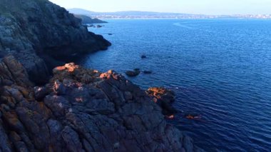 Rocky Coastal Cliff, Sunset Over Blue Ocean 'da. Günbatımı engebeli kıyı kayalarını aydınlatmakta ve derin mavi okyanusla buluşmaktadır. Berrak bir gökyüzünün altında uzak tepeleri olan huzurlu deniz manzarası.