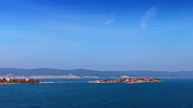 Mavi Deniz ve Dağlık Ufuk ile Sunlit Adası Şehri. Uzak sıradağlar ve sakin deniz manzaralı bir Akdeniz manzarası yaratır..