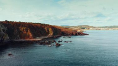 Crimson Rocky Cliffs Overlooking Calm Sea. Scenic coastal landscape with dramatic reddish cliffs, calm turquoise waters, and scattered rocks.