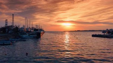 Batan güneş deniz manzarasının engebeli yüzeyine yansıyor. Turuncu gökyüzü gün batımında bulutlarla kaplıdır. Rıhtımda teknelerle çekilmiş görüntüler..