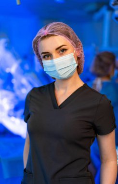 Bir sağlık çalışanı hastane odasında duruyor. Maske takıyor ve saç filesi takıyor. Tıbbi bir prosedüre hazırlanırken etrafını parlak mavi ışıklar sarıyor..