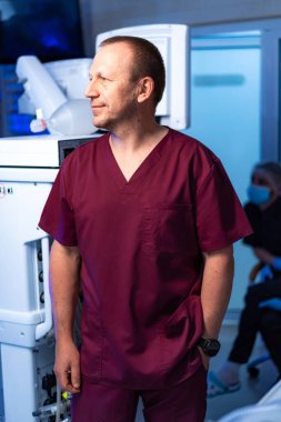 Bordo önlüklü bir sağlık çalışanı tıbbi bir tesiste duruyor. Hasta bakımı hakkında konuşan başka birine bakıyor..