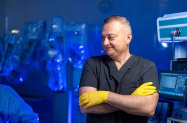 Sağlık hizmetleri ortamında kolları kavuşturulmuş, eldiven takan bir tıp uzmanı. Arka planda cerrahi ekipman ve tıbbi cihazlar görünüyor..