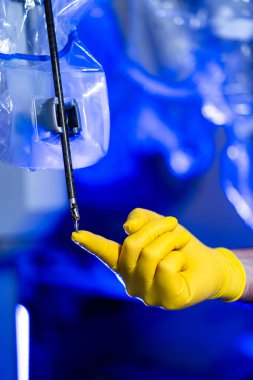 Sarı eldiven takan bir el laboratuvardaki cihazı çalıştırmak için uzanıyor. Ayarda mavi ışık ve temiz ekipmanlar var..