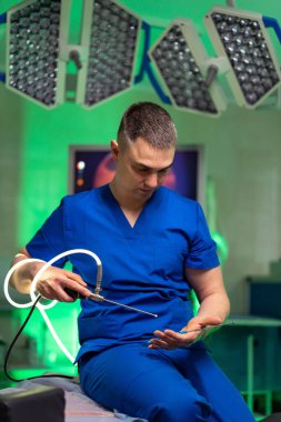 Mavi önlüklü bir tıp uzmanı ameliyathanede oturuyor. Cerrahi bir alet tutuyor ve ameliyata başlamadan önce eline odaklanmış gibi görünüyor..