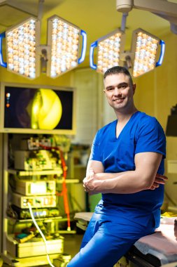 Bir cerrah ameliyat odasında mavi önlük giyerek duruyor. Cerrahi ışıklar yukarıda parlıyor. Odanın etrafına tıbbi ekipmanlar kuruldu. Atmosfer prosedüre hazır olduğunu gösteriyor..