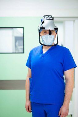 Bir sağlık çalışanı hastane odasında mavi önlük, maske ve yüz maskesi takıyor. Hastane temiz ve düzenli görünüyor. Hastalar yakınlarda..