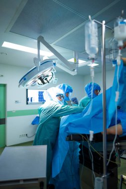 Bir grup tıbbi personel bir hastane ameliyathanesinde birlikte çalışıyor. Ameliyat sırasında önlük, maske ve eldiven takarlar. Odada tıbbi malzemeler ve malzemeler var..