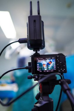 Bir tıbbi ekip ameliyata alınırken, bir tripodun üzerindeki kamera prosedürü yakalıyor. Ekipmanlar ameliyathanede görülüyor. Hasta bakımına odaklanmış durumda..