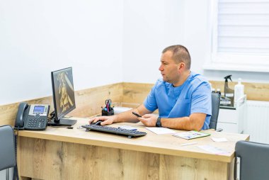 Masada oturan erkek doktor elinde akıllı bir telefonla röntgen ekranına bakıyor. Profesyonel bir erkek doktor bir klinikte masada oturuyor, elinde akıllı bir telefon tutuyor ve muayene sırasında bilgisayar ekranında insan omurgasının 3 boyutlu röntgen modelini analiz ediyor..