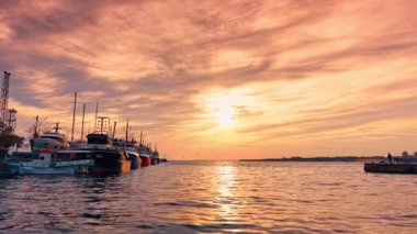 Batan güneşin aydınlattığı engebeli su manzarası. Turuncu gökyüzü bulutlarla kaplı. Suyun üzerinde duran tekneler boyunca uçmak.