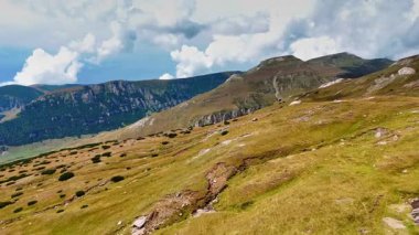 İnsanlar bulutlu bir gökyüzünün altındaki kayalık dağlarla çevrili çimenli tepelerde yürüyor. Çevrede yeşil çalılar ve kahverengi toprak bulunur, doğal bir ayarı gösterir..