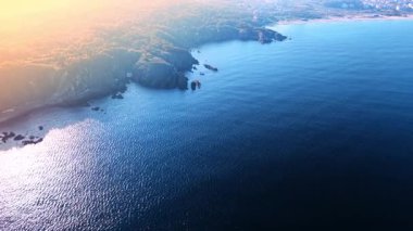 Dalgalar kayalık kıyılara hafifçe çarpar. Güneş ufuktan batıyor, su ve kara üzerine turuncu ve sarı ışık saçıyor. Sahne huzurlu bir kıyı bölgesini gösteriyor..