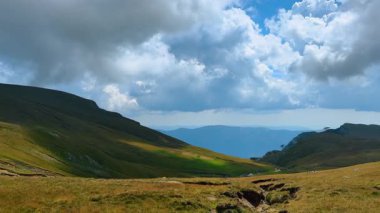 Romanya 'da dağlarla çevrili geniş yeşil bir vadi üzerinde bulutlar toplanıyor. Manzara, parlak bir gökyüzünün altında yuvarlanan tepeleri ve uzak tepeleri barındırır..