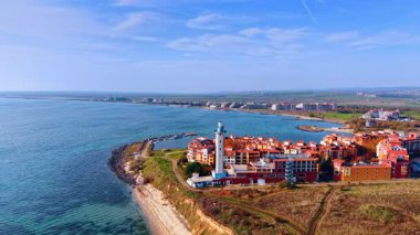 Kıyıda renkli binaların yanında bir deniz feneri duruyor. Okyanus berrak bir gökyüzünün altında uzanıyor. Tekneler rıhtıma yakın, tepeler uzakta..