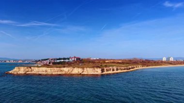 Bir şehrin yakınında küçük bir ada suyun içinde duruyor. Binalar adada ve kıyı şeridi boyunca görülebilir. Gökyüzü birkaç bulutla temiz. Manzara gündüz çekiliyor..