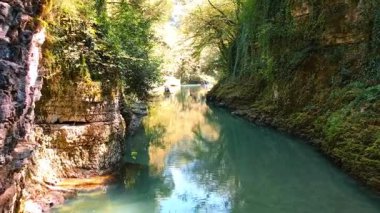 Küçük bir bottaki bir grup insan yüksek kaya duvarları ve yemyeşil ağaçlarla çevrili dar bir su yolunda yavaşça ilerliyorlar. Güneş ışıl ışıl parlıyor ve suyu yansıtıyor.