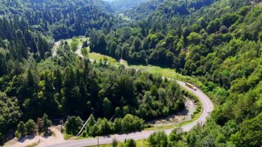 Dönek bir yol, dağlık bir bölgedeki yemyeşil bir araziyi kesiyor. Bölgeyi çevreleyen ağaçlar ve yakınlarda bir nehir akıyor. Gökyüzü açık güneşli bir gün..