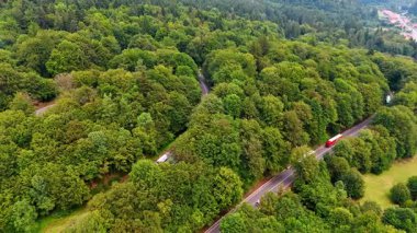 Bir kamyon, kalın yeşil ağaçlarla çevrili dolambaçlı bir yol boyunca gidiyor. Yol uzak bir kasabaya doğru inen ormanlık bir bölgeden geçiyor..