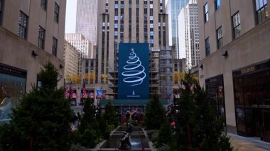 New York, ABD, 18 Aralık 2025: Çeşmenin etrafında güzel yeşil köknar ağaçları yetişiyor. Arka planda rüzgarda dalgalanan Amerikan bayrakları grubu. New York şehir merkezindeki gökdelenin alçak açılı görüntüsü, ABD.