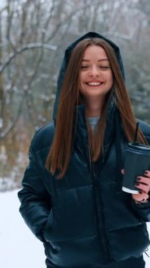 Uzun esmer saçlı mutlu, rahat kadın kışın parkın etrafında yürüyor. Elinde kahve fincanı olan gülümseyen bayan. Dikey video